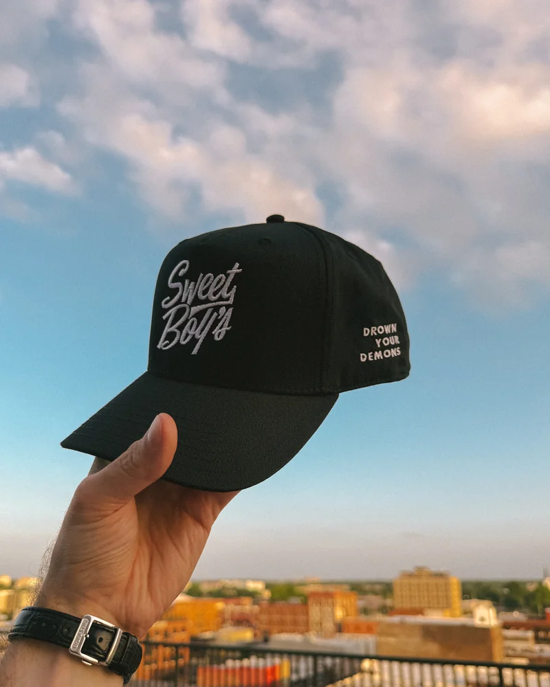 A black Sweet Boy's cap held against a pastel evening sky, with white logo embroidery and Drown Your Demons stitched on the side.
